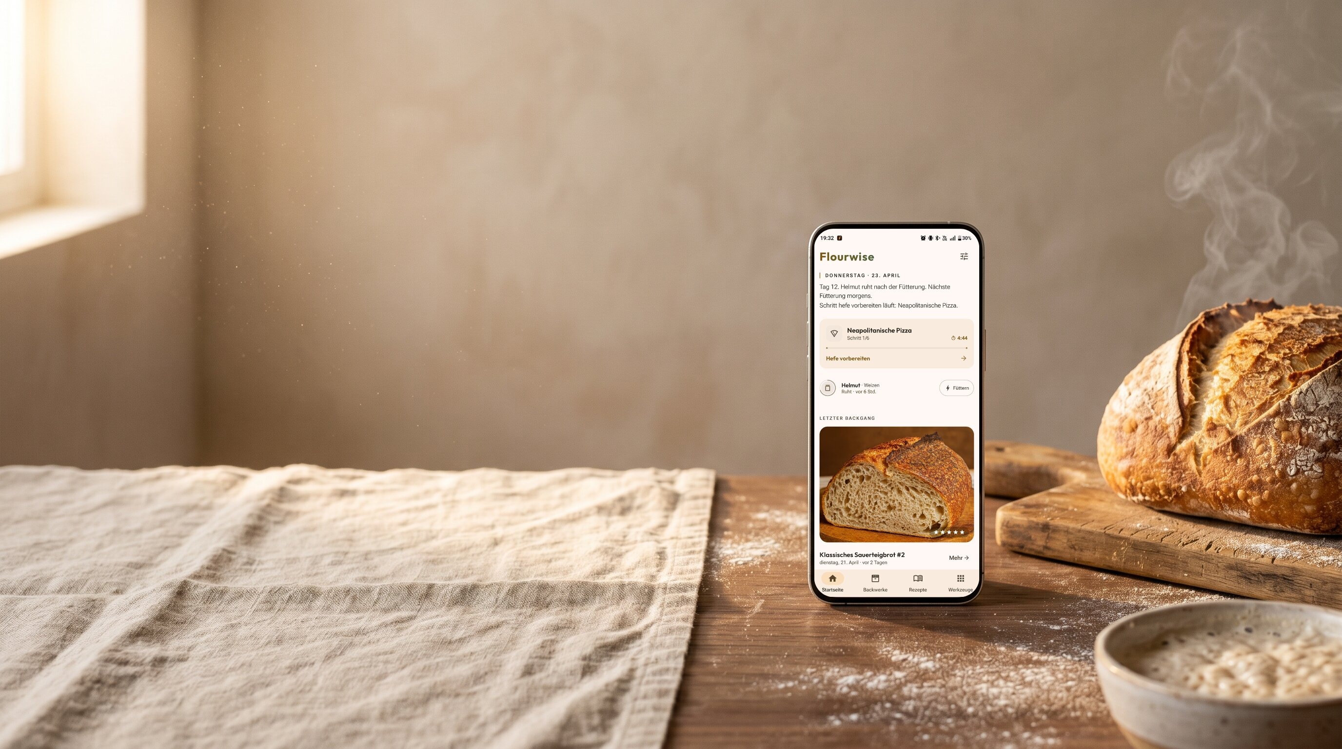 Sauerteigbrot kühlt neben einem Smartphone mit der Flourwise-App ab: aktiver Neapolitanische-Pizza-Timer, Sauerteig-Status und letzte Backsession. Leinentuch, Mehl und Sauerteig-Schüssel auf rustikaler Küchenarbeitsplatte.