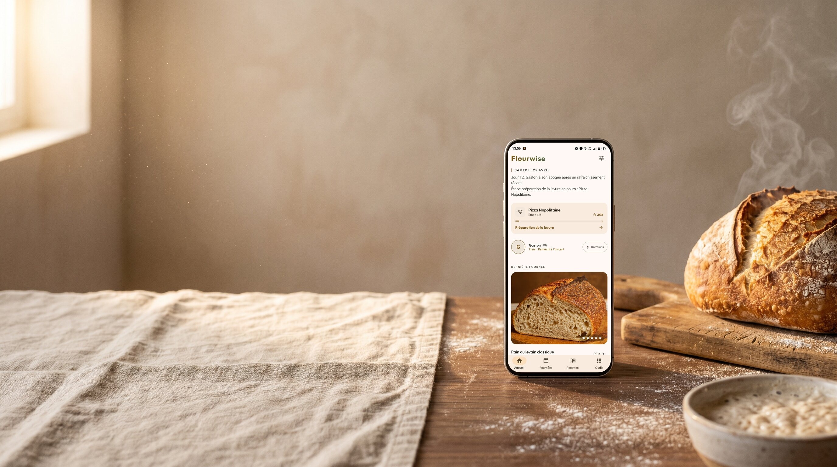 Un pain au levain refroidit à côté d'un téléphone affichant l'application Flourwise : minuteur actif pour pizza napolitaine, état du levain et historique de la dernière fournée. Tissu de lin, farine et bol de levain sur un plan de travail rustique.