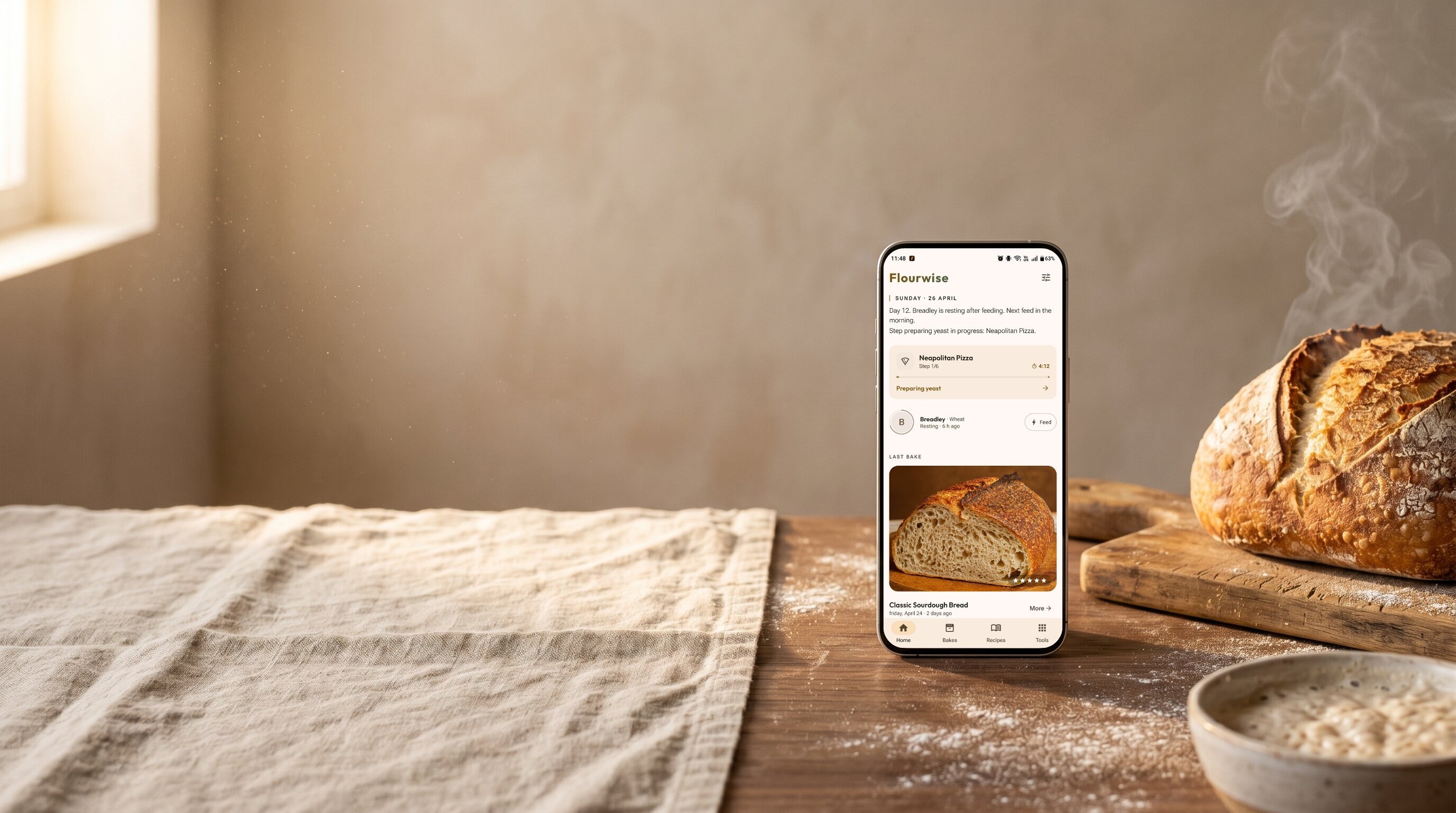 Sourdough loaf cooling next to a phone showing the Flourwise baking companion app: an active Neapolitan pizza timer, sourdough starter status, and the last bake history. Linen cloth, flour and a starter bowl on a rustic kitchen counter.