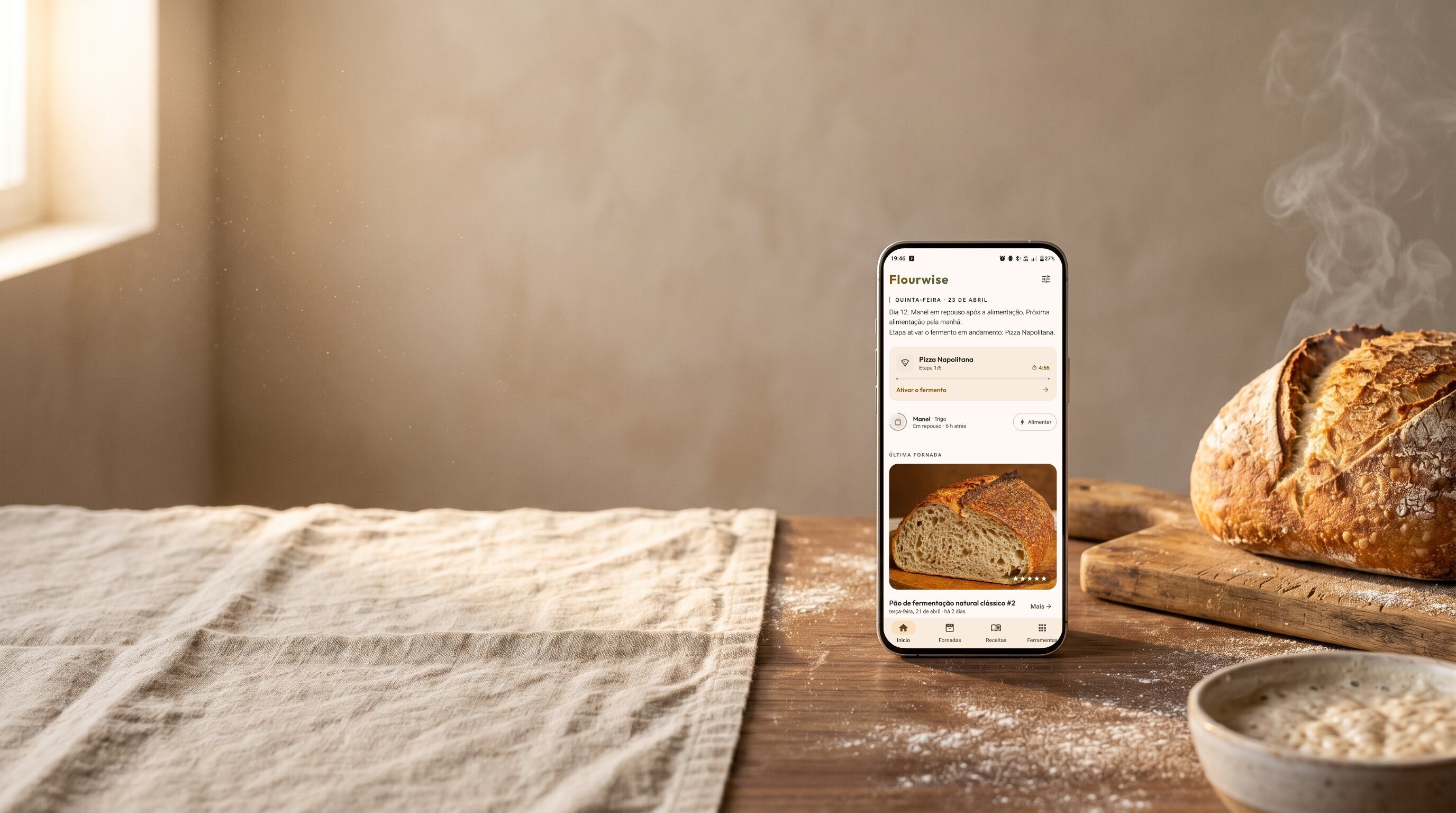 Pão de fermentação natural esfriando ao lado de um celular mostrando o app Flourwise: cronômetro ativo de pizza napolitana, status do fermento natural e histórico da última fornada. Pano de linho, farinha e tigela de fermento sobre uma bancada rústica.